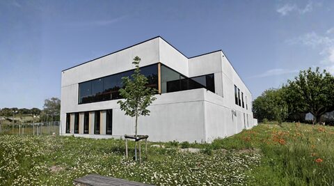 Het bedrijfspand van Maximum Image: een modern grijs gebouw met plat dak en grote donkere raampartijen, gezien vanuit een bloemenweide met margrieten en klaprozen.