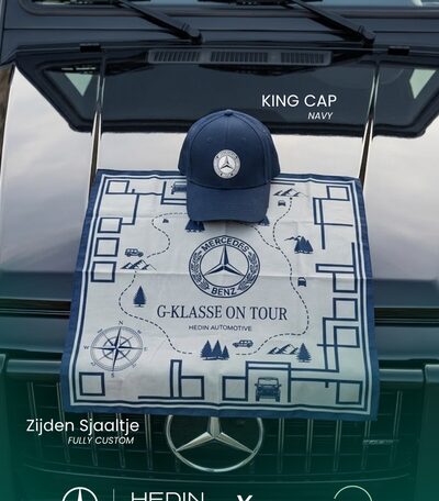Een marineblauwe pet met Mercedes-Benz-logo op een zijden sjaal met het routeontwerp 'G-Klasse On Tour — Hedin Automotive', uitgestald op de groene achterklep van een G-Klasse.