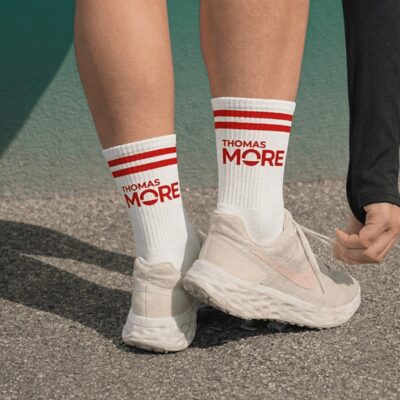 Twee benen in witte sportkousen met rode strepen en het rode logo van Thomas More, boven lichte sneakers op asfalt.